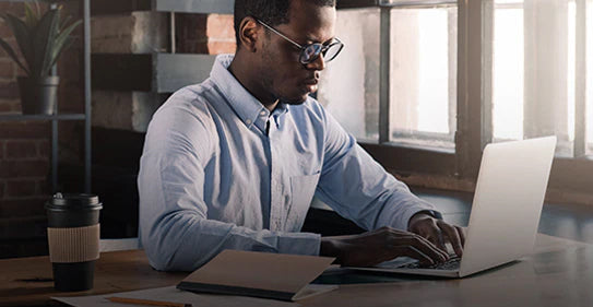 A man wearing glasses and a blue shirt working on a laptop at a desk